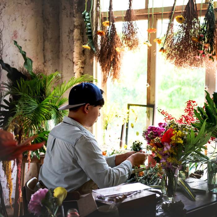 Florist crafting a bouquet of anniversary flowers in a rustic workshop surrounded by fresh blooms and hanging plants.