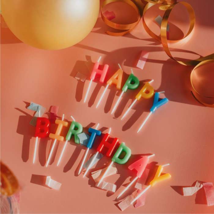 Colorful Happy Birthday candles arranged on a festive orange background with confetti, balloons, and ribbon decorations, representing Happy Birthday Gifts.