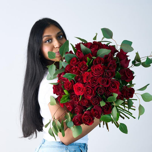 Woman holding a large bouquet of 50 red roses with lush greenery, facing the camera and smiling softly.