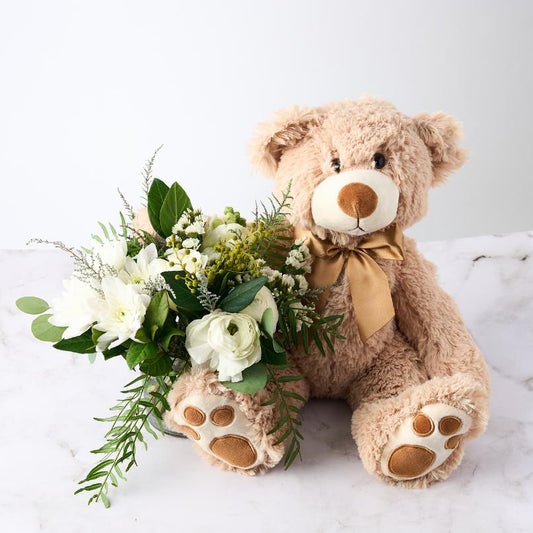 A soft teddy bear with a gold ribbon bow, sitting beside a bouquet of white flowers and greenery on a white marble surface.