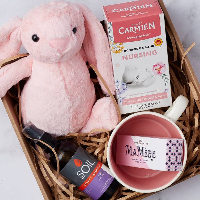 Close-up of Blissful Mum & Baby Set showing a pink plush bunny, nursing tea, SOiL organic baby oil, a ceramic cup, and almond cranberry nougat.