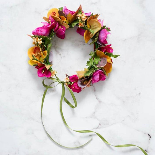 Colorful flower crown made with pink, orange, and yellow flowers, accented with green leaves and a green ribbon, arranged on a marble surface.