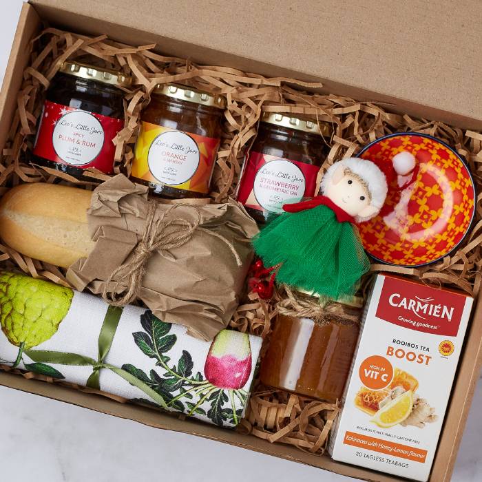 Breakfast with Santa Gift Box featuring artisanal jams, Carmién Rooibos Boost Tea, honey, a bread roll, a festive doll, a colourful ceramic dish, and a beautifully designed tea towel, all presented in a kraft box with natural filler.