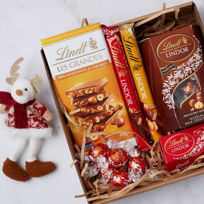 Festive gift box featuring an assortment of Lindt chocolates, including milk chocolate bars, truffles, and hazelnut selections, alongside a decorative plush reindeer figurine.