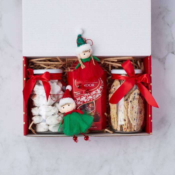 Christmas-themed gift box featuring jars of white meringues, biscotti, and Lindt hot chocolate, adorned with festive red ribbons and two adorable elf decorations.