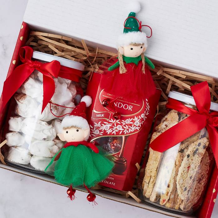 Holiday gift box showcasing jars of meringues and biscotti, a Lindt hot chocolate tin, and two cute elf dolls, wrapped in festive packaging.