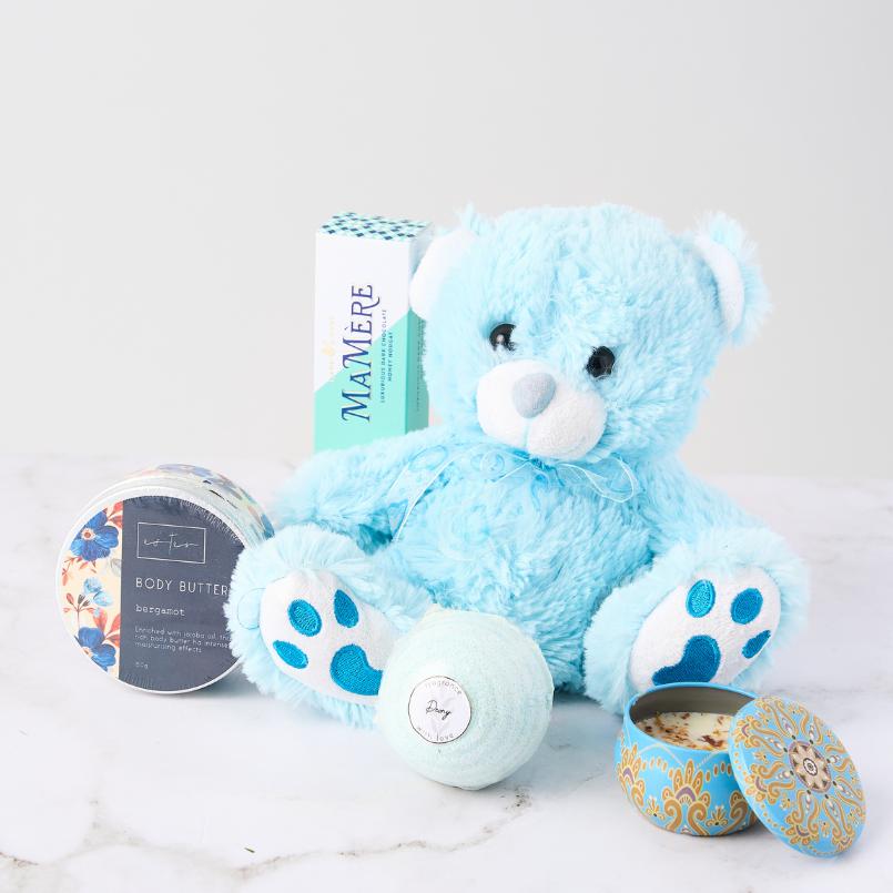 Cosy Cuddles Gift Set from Flower Guy, featuring a plush blue teddy bear, Mamère confection, bergamot body butter, bath bomb, and a decorative tin candle.