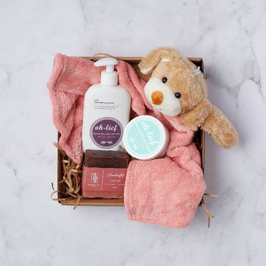 Top view of the Cuddle and Care Baby Gift Set, featuring a plush bear, Oh-Lief natural olive baby shampoo, bum balm, a pink baby blanket, and a handcrafted soap bar, elegantly packaged in a kraft gift box with shredded paper.