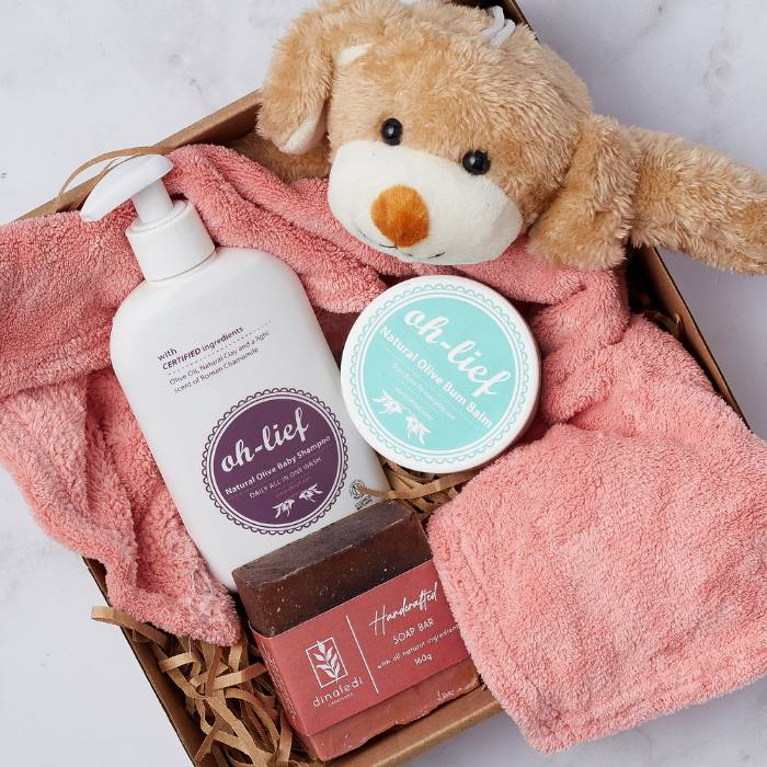 Close-up of the Cuddle and Care Baby Gift Set contents, including a soft plush bear, Oh-Lief baby shampoo and bum balm, a pink baby blanket, and a handcrafted soap bar, displayed in a kraft box with natural packaging.