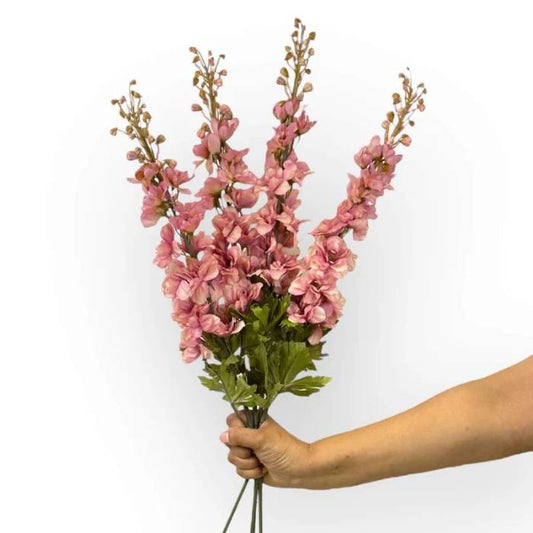 A hand holding a bouquet of dusty pink Delphinium artificial flowers with green leaves, showcasing the elegant, realistic design of the stems