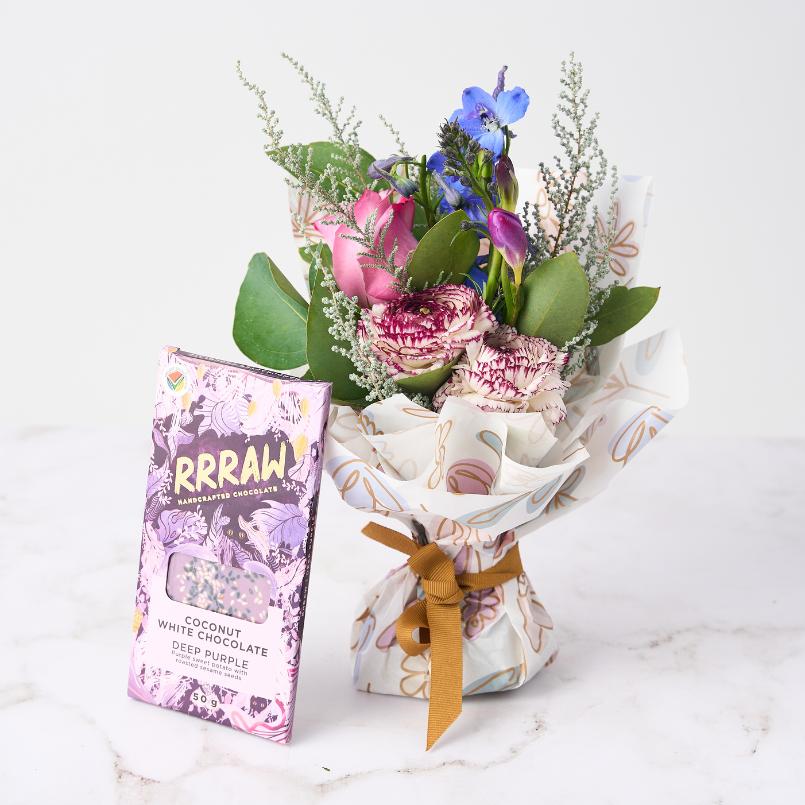 Floral Fantasy Posy featuring pink roses, purple ranunculus, blue delphiniums, and greenery, paired with RRRAW Coconut White Chocolate Deep Purple, elegantly wrapped and ready for gifting.