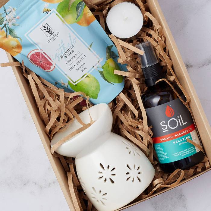 Close-up of the Calm and Peaceful Spa Gift Box showcasing grapefruit & lime bath salts, a ceramic oil burner, a tealight candle, and SOiL organic relaxing massage oil, arranged with wood wool packaging for a cozy presentation.