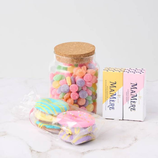 A jar of multicoloured jelly tots with a cork lid, two Mamère nougat bars in pink and yellow packaging, and a pack of pastel-coloured biscuits, neatly arranged on a white marble surface.