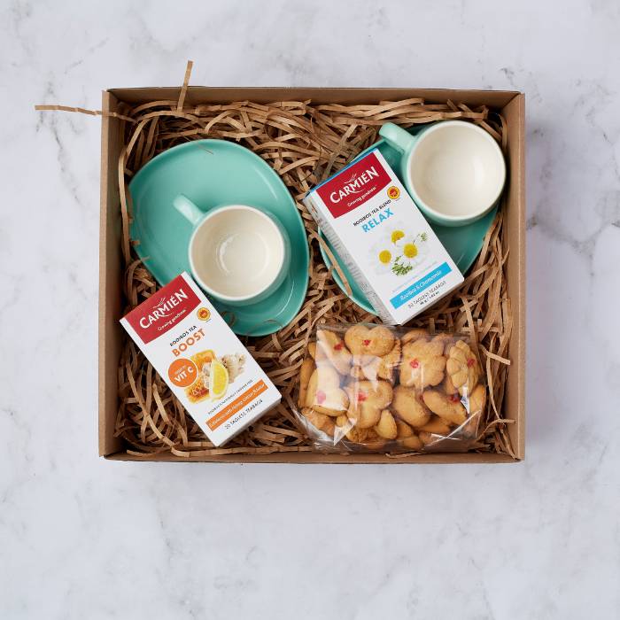 A beautifully packaged Tea Remedy Gift Box containing Carmién Rooibos Tea, mint green ceramic cups with saucers, and freshly baked biscuits, displayed on a natural bed of straw.