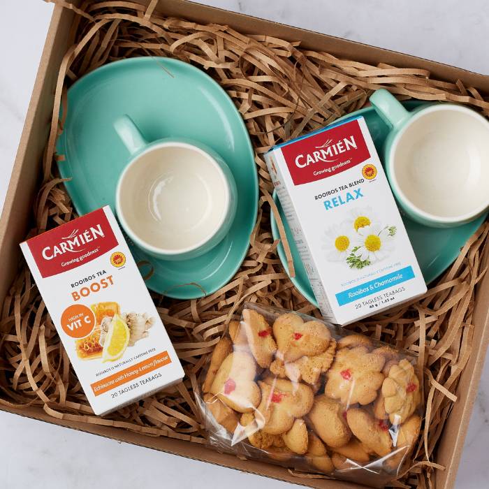 Close-up of the Tea Remedy Gift Box featuring two boxes of Carmién Rooibos Tea, elegant mint green ceramic tea cups with saucers, and a bag of heart-shaped biscuits, perfect for gifting or personal relaxation.