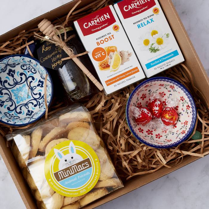 Close-up of Tea Time Treat gift box featuring Carmien Rooibos tea, honey jar with wooden dipper, Lindor chocolates, vanilla biscuits, and two handcrafted ceramic bowls in vibrant patterns.